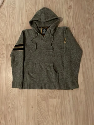 Grå hoodie från Gant i lammull - Mysig grå hoodie från Gant i 100% lammull med broderad Gant-logga och New Haven-märke på bröstet. Huva med dragsko, ribbade muddar och ficka framtill. Snygga svarta och gula detaljer på ärmen för extra stil.