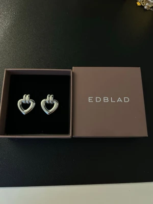 Silverfärgade hjärtörhängen edblad - Säljer ett par stilrena örhängen från EDBLAD i silverfärgad metall. Örhängena har en öppen hjärtformad design och stift på baksidan. 