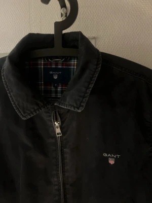 Svart jacka från GANT - Klassisk svart bomberjacka från GANT med dragkedja framtill och broderad logga på bröstet. Jackan har rutigt innerfoder och två fickor på sidorna. Perfekt för dig som gillar stilren och tidlös design.