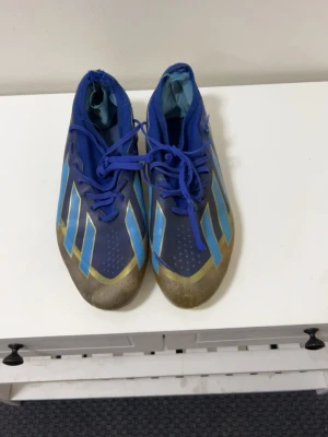 Blå fotbollsskor med gula detaljer - Snygga fotbollsskor i blått med gula och ljusblå detaljer. Skorna har snörning och en låg profil med rund tå. Yttersulan har dobbar för bra grepp på planen. Perfekta för dig som vill sticka ut på fotbollsplanen.