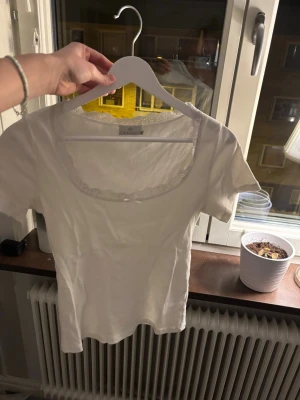 Vit ribbad topp med spets och rosett - Söt vit ribbad topp med korta ärmar, spetskant och liten rosett vid urringningen. Perfekt att styla med jeans eller kjol för en trendig look.