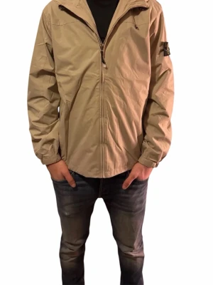Beige vindjacka från Stone Island - Säljer en beige vindjacka från Stone Island med huva och dragkedja framtill. Jackan har den klassiska Stone Island-loggan på vänster ärm och är tillverkad i lätt polyester. Perfekt för dig som vill ha en clean och stilren look.