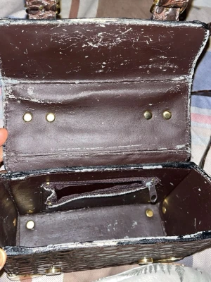 Brun handväska i skinn med dragkedja - Snygg brun handväska i skinn med rektangulär form och guldfärgade nitar. Väskan har ett innerfack med dragkedja och lock som stängs med tryckknappar. Perfekt för att bära det viktigaste och ger en klassisk look. Det är några tecken på användning men syns inte på utsidan bara insidan 