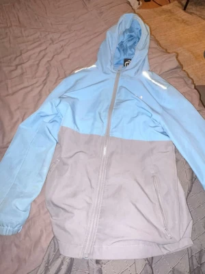 Blå och grå vindjacka med huva - Snygg tvåfärgad vindjacka i ljusblått och grått med huva och hel dragkedja framtill. Jackan har långa ärmar, två sidofickor med dragkedja och elastiska muddar vid ärmsluten. Perfekt för blåsiga dagar.