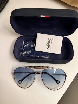 Tommy Hilfiger solglasögon  - Snygga aviator solglasögon från Tommy Hilfiger  med metallbågar i silver och blåtonade glas. Översta delen av bågen har ett sköldpaddsmönstrat inslag. Kommer med originalfodral och putsduk. Perfekt accessoar för en cool och trendig look.