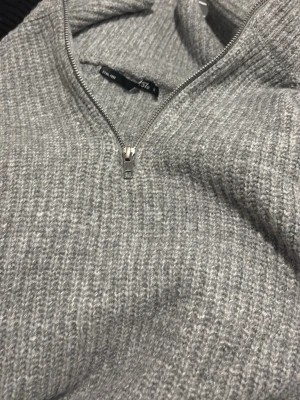 Grå stickad halvzip-tröja från lager - Säljer en grå stickad tröja från H&M med halvzip och ribbad struktur. Tröjan har hög krage och är perfekt för lager på lager. Enkel och stilren design som passar till det mesta.