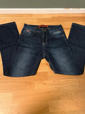 Mörkblå Hilfiger Denim jeans - Säljer ett par mörkblå jeans från Hilfiger Denim med klassisk fyrficksmodell och snygga kontrastsömmar. Jeansen har raka ben och subtila slitningar framtill för en avslappnad look. Perfekta till vardags för en stilren outfit.