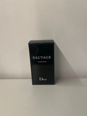 Dior Sauvage Eau de Parfum - Dior Sauvage Eau de Parfum i en elegant svart rektangulär kartong med vit text. Parfymen är känd för sin fräscha och maskulina doft och kommer i en stilren förpackning som utstrålar lyx och modernitet. Perfekt för dig som gillar exklusiva dofter.