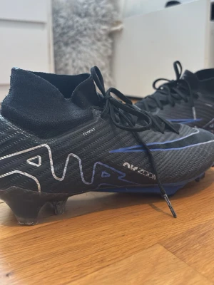 Nike Mercurial Zoom fotbollsskor svart/blå - Säljer ett par Nike Mercurial Zoom fotbollsskor i svart med blå och vita detaljer. Skorna har snörning, hög strumpliknande krage och är tillverkade i syntetmaterial med textil. Perfekta för dig som vill ha grym touch och stil på planen.