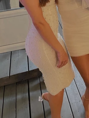 Oh polly pärlklänning - Säljer en elegant vit kort klänning med tunna axelband och pärldetaljer över hela tyget. Klänningen har en figurnära passform och en lätt draperad urringning. Perfekt för dig som vill sticka ut med en unik och stilren look. Perfekt till studenten 