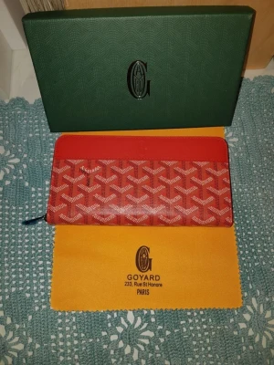 Röd Goyard plånbok med mönster - Snygg röd plånbok från Goyard med ikoniskt vitt mönster och dragkedja runtom. Plånboken har en avlång form och klassisk logga på framsidan. Perfekt för dig som vill ha något både stilrent och trendigt.