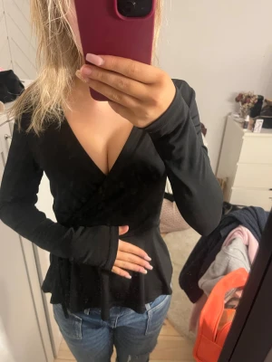 Svart omlottopp med v-ringning - Snygg svart omlottopp med djup v-ringning och långa ärmar. Toppen har en markerad midja och volang nedtill som ger en feminin siluett. Perfekt att styla med jeans eller kjol för en trendig look.