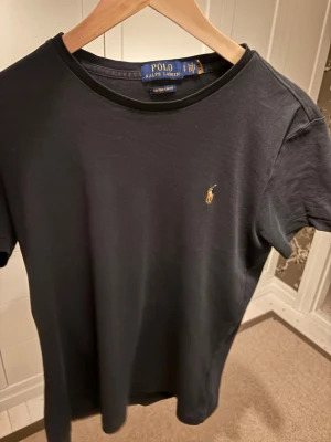 Svart t-shirt från Polo Ralph Lauren - Klassisk svart t-shirt från Polo Ralph Lauren med rund halsringning och liten broderad logga på bröstet. T-shirten har korta ärmar och är tillverkad i mjuk bomull för en skön känsla. Perfekt till jeans eller shorts. Funkar även för Storlek M. 
