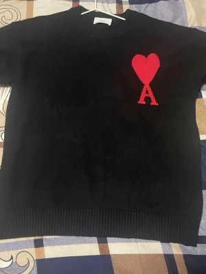 Svart stickad tröja från Ami Paris - Svart stickad tröja från Ami Paris med B röd hjärtlogga och bokstaven A på bröstet. Tröjan har rund halsringning och ribbstickade muddar vid ärmslut och nederkant. Perfekt för dig som gillar stilrena plagg med en twist.