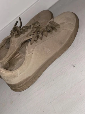 Beige sneakers från Polo Ralph Lauren - Stilrena beige sneakers från Polo Ralph Lauren med klassisk låg profil och rund tå. Skorna har snörning och är tillverkade i mocka med diskret logga på sidan. Perfekta för dig som gillar en clean och enkel look.