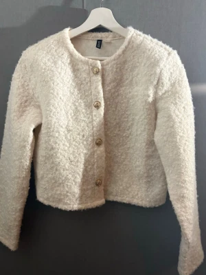 Vit bouclékofta från H&M - Stilren vit bouclékofta från H&M med rund halsringning och fyra dekorativa guldfärgade knappar framtill. Koftan har lång ärm och en mjuk, fluffig struktur som ger en trendig look. Perfekt att matcha med jeans eller kjol för en chic outfit.