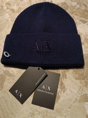 Mörkblå mössa från Armani Exchange - Snygg mörkblå ribbstickad mössa från Armani Exchange med uppvikt kant och broderad A|X-logga framtill. Tillverkad i 100% bomull för skön känsla och stilren look. Perfekt accessoar för kalla dagar.