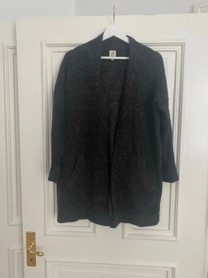 Mörkgrå lång kofta från Ateljé - Mysig och stilren mörkgrå lång kofta från Ateljé. Koftan har öppen front, två fickor framtill och är tillverkad i ett mjukt, lite luddigt material. Perfekt för lager på lager och enkel att matcha med olika outfits.