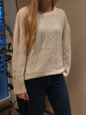 Vit kabelstickad tröja från 157 - Mysig vit stickad tröja från 157 med kabelstickat mönster framtill och diskreta diamantformer på sidorna. Tröjan har rund halsringning och ribbade muddar vid ärmslut och nederkant. Perfekt för kalla dagar och enkel att matcha.