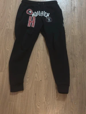 Svarta mjukisbyxor med patchar - Svarta mjukisbyxor med flera patchar och tryck framtill, bland annat ett stort rött H och texten 'RICH'. Byxorna har snörning i midjan och muddar vid bensluten för en skön och avslappnad look.