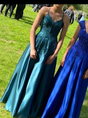 Grön balklänning med spetsdetaljer - Grön långklänning med spets upptill och snörning i ryggen. Klänningen har smala axelband och en utsvängd kjol, perfekt för speciella tillfällen som exempelvis bal. Klänningen är köpt på JJ’s House för nypris ca. 2 200kr.