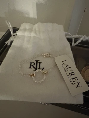 Silverarmband från Ralph Lauren - Snyggt och stilrent armband från Ralph Lauren med tunn silverkedja och en tvinnad cirkel i mitten. Armbandet har små guldfärgade detaljer som ger en lyxig touch. Perfekt accessoar för dig som gillar minimalistisk design och exklusiva märken.