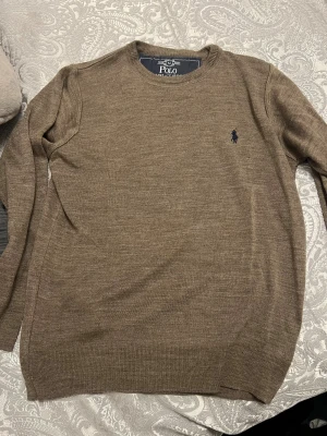 Brun stickad tröja från Polo Ralph Lauren - Snygg brun stickad tröja från Polo Ralph Lauren med klassisk rund hals och diskret broderad logga i mörkblått på bröstet. Tröjan har långärmad passform och är tillverkad i mjukt material som känns skönt mot huden.