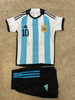 Argentina Messi fotbollströja Adidas - Säljer en klassisk Argentina fotbollströja från Adidas med Messi 10-tryck. Tröjan är kortärmad, har blå och vita vertikala ränder, svarta detaljer och AFA-emblem på bröstet. Materialet är lätt och andas, perfekt för fotbollsträning eller match.