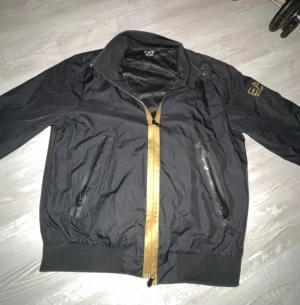 Svart vindjacka EA7 Emporio Armani - Svart vindjacka från EA7 Emporio Armani med guldiga detaljer och dragkedja. Jackan har två snedställda fickor med dragkedja, ribbad mudd vid midja och ärmslut samt tryckt logga på ärmen.