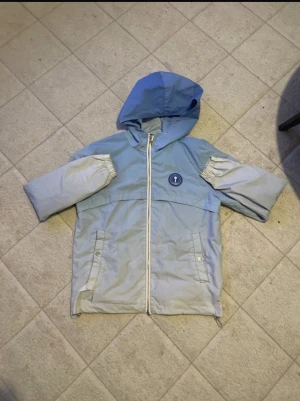 Blå vindjacka från Trapstar - Säljer en blå och ljusgrå vindjacka från Trapstar med huva och dragkedja framtill. Jackan har broderad Trapstar-text i blått på ryggen och ett runt blått märke med guldtext på bröstet. Fickor med knappstängning och elastiska ärmslut. Perfekt för dig som gillar streetwear.