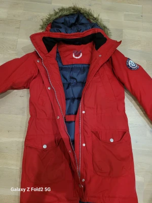 Röd parkas från Etirel med päls - Parkas från Etirel i rött med fuskpäls på huvan och snygga detaljer som stora fickor med knappar och dragkedja framtill. Jackan har ett campus sportswear-märke på ärmen och är fodrad med mörkgrått innerfoder. Perfekt för kalla dagar.