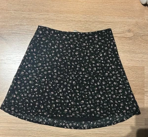 Svart blommig kjol från H&M Divided - Supersöt svart kortkjol från H&M Divided i storlek 32 med små blommor i rosa och vitt, har en dragkedja på sidan!
