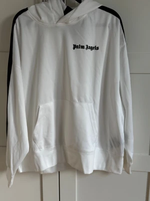 Vit hoodie med svarta ränder - Säljer en vit hoodie från Palm Angels med svarta broderade loggan på bröstet och svarta ränder längs ärmarna. Tröjan har huva, känguruficka och är långärmad. Materialet är mjukt och känns som bomull eller polyester, perfekt för chill dagar.