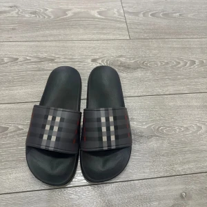 Svarta burberry tofflor  - Svarta sandaler med bred rem i klassiskt rutigt mönster i grått, svart och rött. Snygg och enkel design med platt sula, perfekt för sommardagar. Remmen har en stilren look som passar till många outfits.