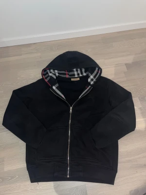 Svart hoodiejacka från Burberry M - Svart hoodiejacka från Burberry i storlek M med klassiskt rutig foder i huvan i svart, vitt, grått och rött. Jackan har dragkedja framtill, två fickor och långärmade ärmar. Perfekt för dig som gillar stilren design med lyxig touch.