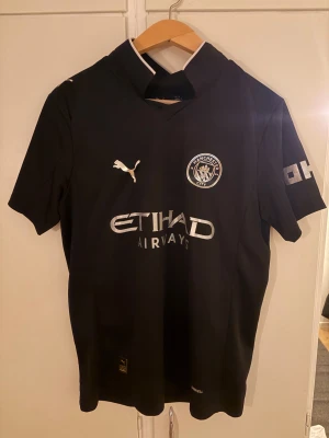 Manchester City svart matchtröja Puma - Säljer en svart Manchester City fotbollströja från Puma med silverdetaljer, klubbmärke och Etihad Airways-tryck på bröstet. Nummer 10 och namnet Cherki på ryggen. Klassisk krage med vita detaljer. Tillverkad i lätt och ventilerande material.