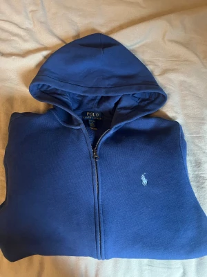 Blå hoodie från Polo Ralph Lauren - Snygg marinblå hoodie från Polo Ralph Lauren med dragkedja och huva. Tröjan har lång ärm, broderad logga på bröstet och är gjord i mjuk bomull. Storleken är L 14-16 år. Hör av er om minsta lilla fråga, priset är inte hugget i sten 