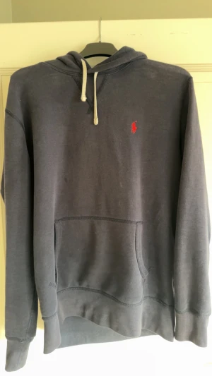 Mörkblå hoodie från Polo Ralph Lauren - Snygg mörkgrå hoodie från Polo Ralph Lauren med klassisk röd broderad logga på bröstet. Tröjan har huva med vit snörning, stor magficka och långa ärmar. Mjuk bomullskänsla och relaxed passform, perfekt för chill dagar.