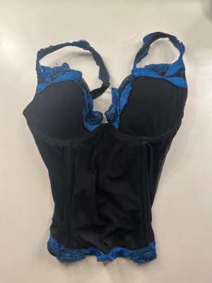 Svart och blå korsett med blommdetaljer - Säljer en svart korsett-topp med blå  blommdetaljer längs kanterna och axelbanden.       Toppen har justerbara axelband och 3 rader med knäppning där bak. Storlek S. Priset är diskuterbart💕