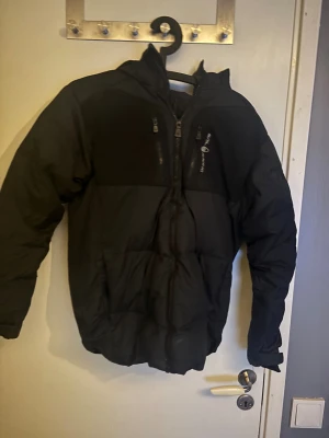 Svart pufferjacka Sail Racing - Svart pufferjacka från Sail Racing med huva och coola detaljer som Sail Antarctica Expedition-märke på ärmen. Jackan har flera dragkedjor framtill och logga på ryggen. Perfekt för kalla dagar med sin tjocka och fluffiga design. Snygg och funktionell för dig som gillar äventyr.