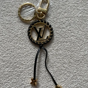  Vuitton  nyckelring i guld och svart - Exklusiv  Vuitton nyckelring i guld- och svartfärgad metall med -logga i mitten. Rund form med flätad svart detalj runt kanten och två svarta band med guldfärgade blommor längst ner. Perfekt accessoar för att lyfta din väska eller nycklar.
