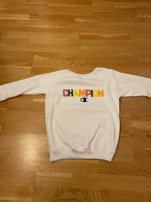 Vit Champion sweatshirt med färgglatt tryck - Vit sweatshirt från Champion med lång ärm och rund hals. Framsidan har ett stort CHAMPION-tryck i rött, gult och orange samt den klassiska Champion-loggan. Tröjan har ribbade muddar vid ärmslut och nederkant, perfekt för en chill och sportig look.