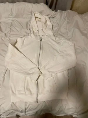 Vit hoodie med dragkedja och huva - Säljer en stilren vit hoodie från 157 med hel dragkedja framtill och praktisk huva med snörning. Tröjan har långa ärmar, ribbade muddar och mjukt bomullsmaterial som är skönt mot huden. Perfekt för chill dagar och enkel att matcha med jeans eller joggers.