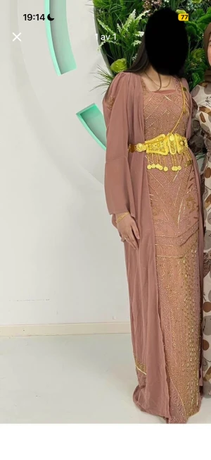 Kurdisk klänning i storlek S/M - Elegant långklänning i ljusrosa med detaljerat guldmönster. Klänningen har långa, lösa ärmar och ett exklusivt, glittrigt tyg som ger en lyxig känsla. Perfekt för dig som vill sticka ut på festen.
