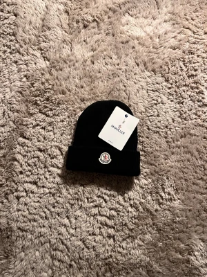 Svart Moncler mössa i ull - Svart mössa från Moncler med klassisk uppvikt kant och Moncler-logga framtill. Stickad i mjuk ull som håller dig varm och ger en clean look. Perfekt för dig som gillar stilrena accessoarer med exklusiv känsla.