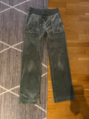 Grönt mjukissett Juicy Couture XXS - Snyggt grönt mjukissett från Juicy Couture i storlek XXS. Byxorna har bred resår i midjan med snörning, stora fickor fram och bak med knappdetaljer. Mjukt och lent material som ger en chill vibe. Byxorna är uppsydda hos skräddare. Innerbenslängd ca 66 cm.