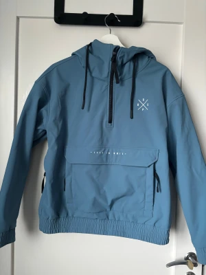 Blå skidjacka från Dope XS - Snygg blå vindjacka från Dope med huva. Jackan har half zip framtill, stor magficka med dragkedja och dragkedjor vid sidan. Insidan är lätt fodrad med mjuk fleece