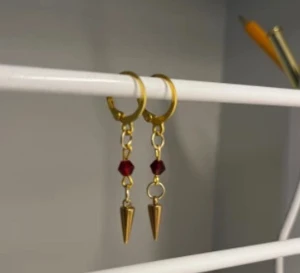 Guldiga örhängen med röda pärlor - Snygga örhängen i guldton med hängande röda pärlor och spetsiga konformade detaljer. Perfekt för dig som vill ha en edgy och unik stil. Passar till både street och trendiga outfits. Rostfritt stål och handgjort