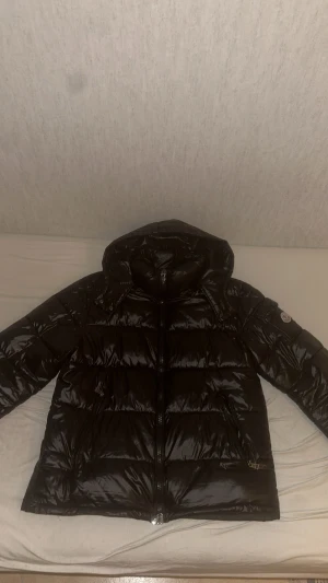 Moncler Maya Jacka Svart - Säljer en svart Moncler Maya Jacka med huva och dragkedja framtill. Jackan har Moncler-logga på ärmen, två fickor med dragkedja och snygga detaljer i metall. Perfekt för kalla dagar och riktigt stilren look. Väldigt bekväm och precis som äkta då de är bra kvalite + NFC skann finns. Saknas en knapp på en hand fast det syns inte ens, väldigt bekväm och skön och passar folk som har storlek M - L. 