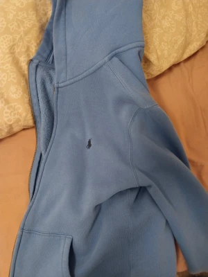 Blå hoodie från Polo Ralph Lauren - Snygg blå hoodie från Polo Ralph Lauren med dragkedja och klassisk broderad logga på bröstet. Tröjan har huva, fickor framtill och är fodrad med mjuk fleece på insidan. Perfekt för chill dagar och enkel att matcha med jeans eller joggers.